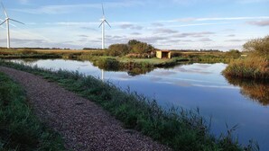 Escena tranquila junto al río con un sendero de grava, un pequeño edificio y aerogeneradores a lo lejos.