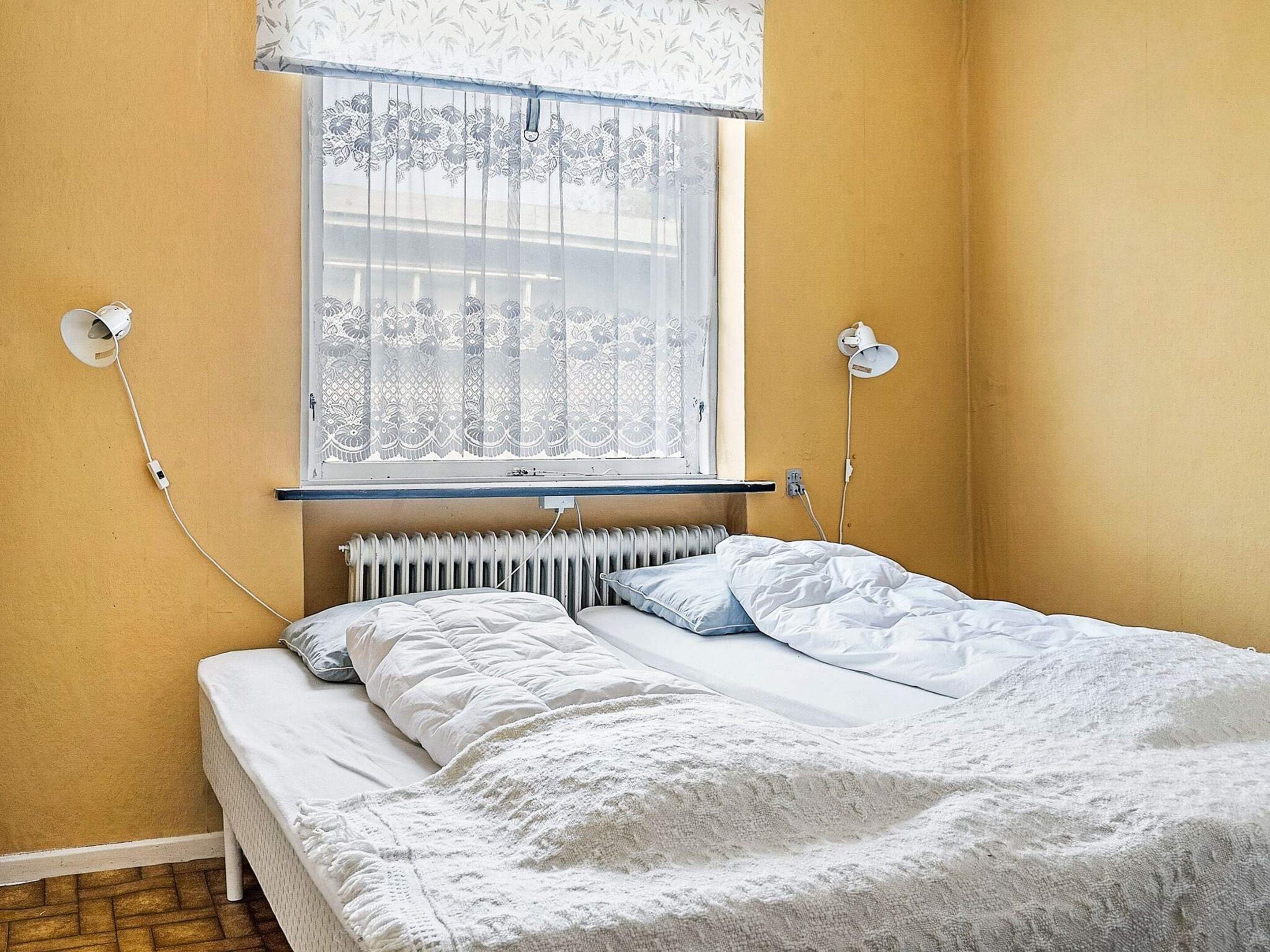 Un dormitorio con cama, un radiador, una ventana con cortina y dos lámparas fijadas a la pared.