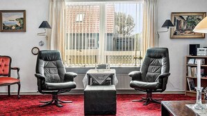 Una habitación con dos sillones de cuero, un pequeño otoman, un radiador, una ventana con cortinas, una estantería y un cuadro enmarcado en la pared.