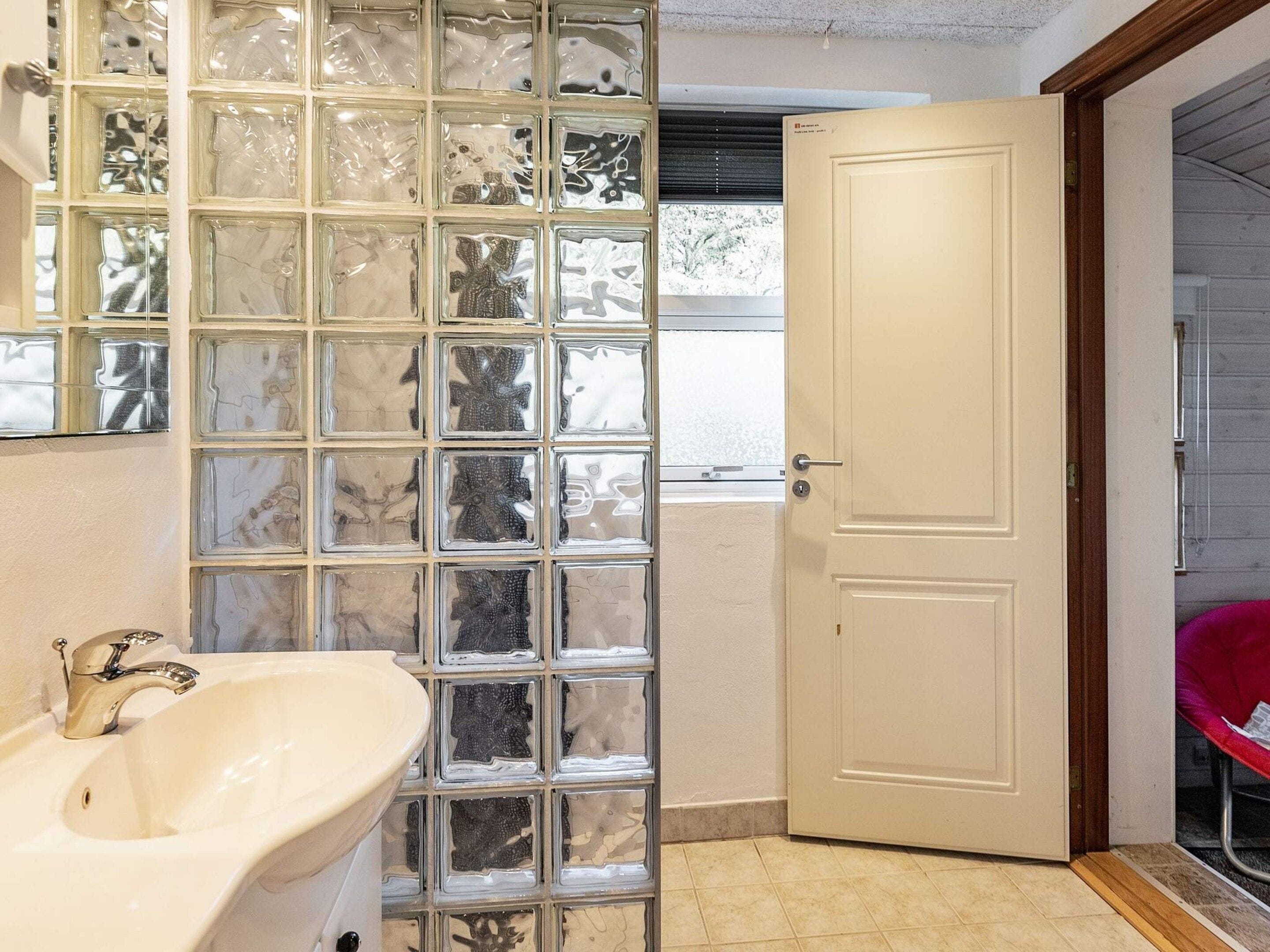 Un baño con lavamanos blanco, separador de vidrio bloqueado, puerta blanca y ventana con persianas.