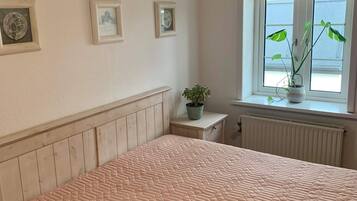 A bedroom with a bed, a nightstand, a window with plants, and framed pictures on the wall.