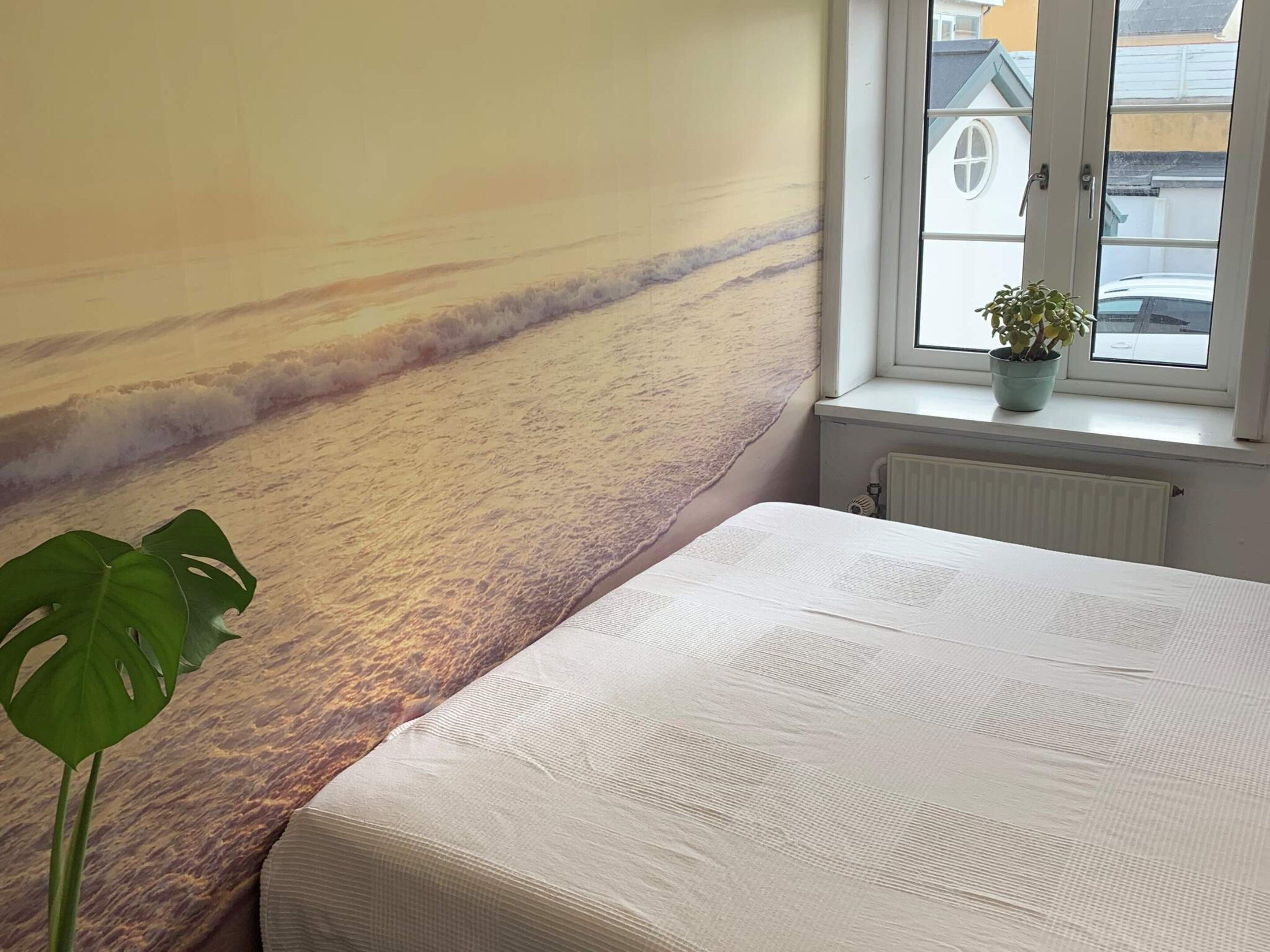 A bedroom with a large wall mural of a beach, a window with a view of buildings, and a potted plant.