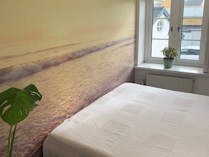 A bedroom with a large wall mural of a beach, a window with a view of buildings, and a potted plant.
