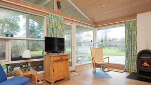 A cozy living room with a television on a wooden cabinet, a fireplace, and large windows offering a view of the outdoors. - Holiday Home in NykÃ¸bing Sj (Nykobing Sjaelland)