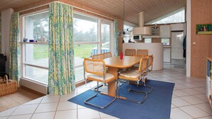 A dining area with a wooden table and wicker chairs, a blue rug, and a view of the outdoors through large windows. - Holiday Home in NykÃ¸bing Sj (Nykobing Sjaelland)