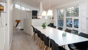 Une cuisine moderne avec des meubles blancs, un îlot central, des luminaires suspendus et un coin repas comprenant une longue table et des chaises.