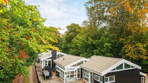 Une maison moderne avec une extension en verre, entourée d’arbres et dotée d’une allée pavée.