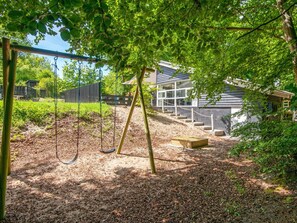A playground with swings and a slide, surrounded by trees and a building with large windows. - 4 Star Holiday Home in Glesborg (Glesborg)