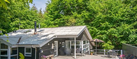 Una cabana rústica amb teulada metàl·lica, un porxo cobert amb seients exteriors i un paisatge forestal al voltant.