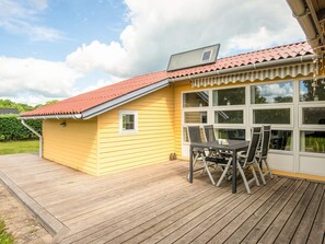 A yellow building with a red roof, a covered patio with a table and chairs, and a large window.