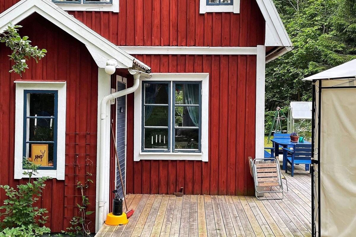 A red wooden house with a covered deck, a window with curtains, and a small table with chairs.