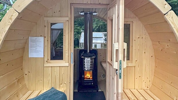 A wooden cabin with a central wood-burning stove, a bench, and a small window. - 4 Person Holiday Home in Karlsborg (Karlsborg)