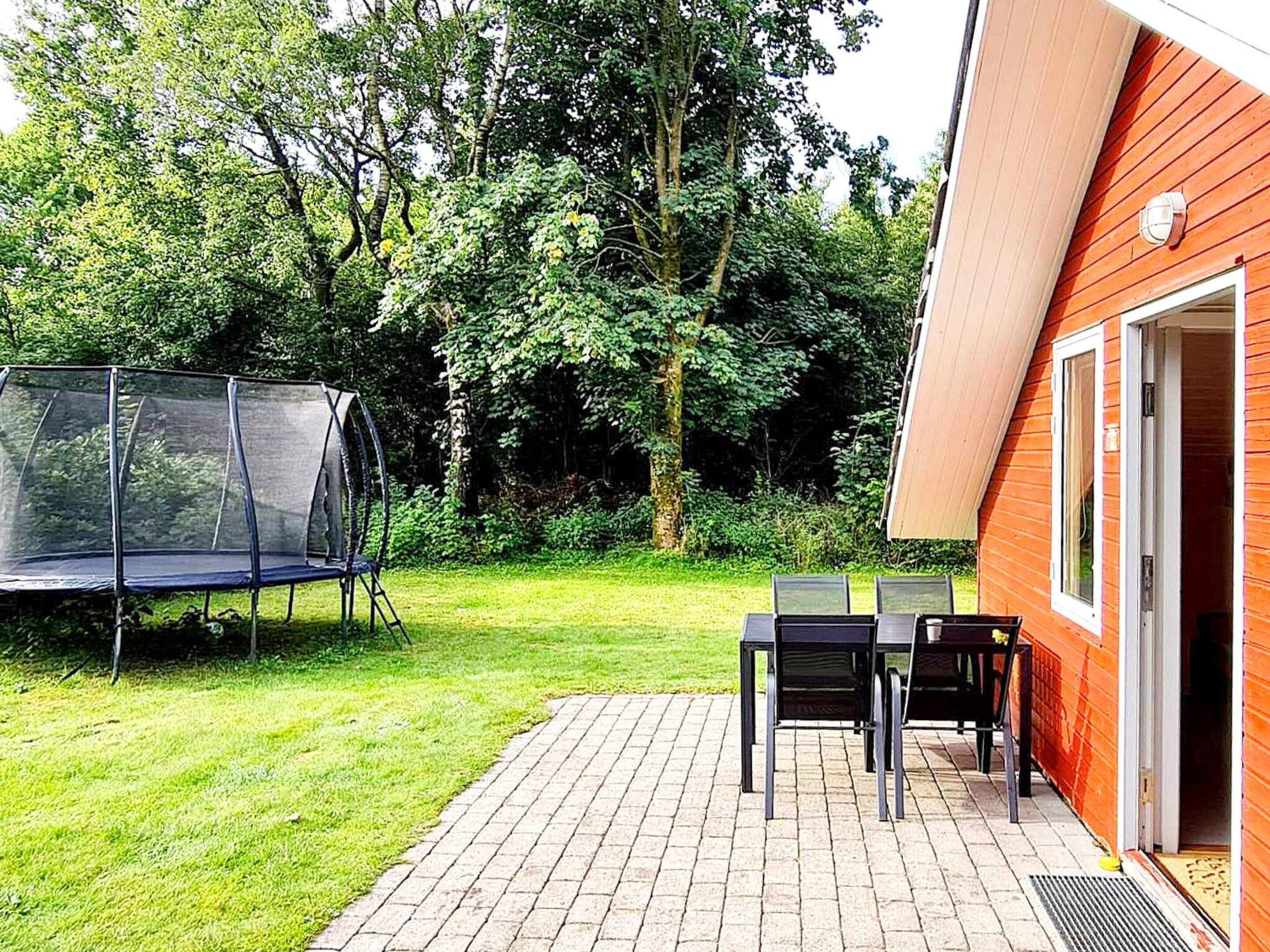 Eine Terrasse mit Trampolin, Tisch und Stühlen, umgeben von einem Holzgebäude und Bäumen.