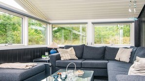 Ein modernes Wohnzimmer mit einem Ecksofa, einem Glastisch und Blick auf Grünflächen durch große Fenster.
