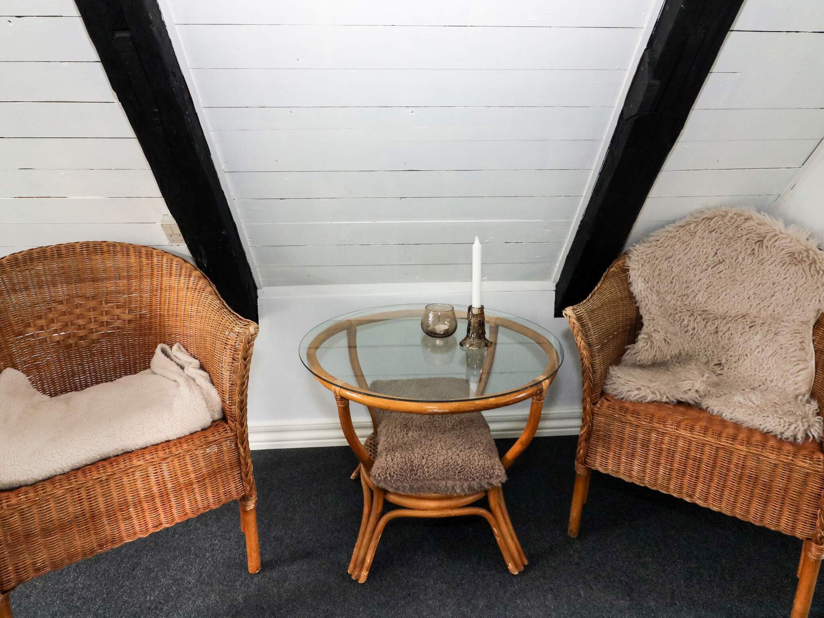 Habitación de esquina con dos sillas de mimbre, una mesa de vidrio y un taburete pequeño.