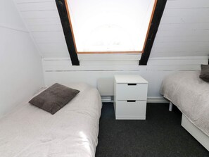 Una habitación pequeña con paredes blancas, dos camas, una mesita de noche blanca y un tragaluz.