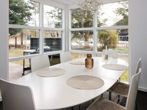 Una mesa de comedor redonda con sillas blancas, una araña y ventanas amplias que dan a un jardín.