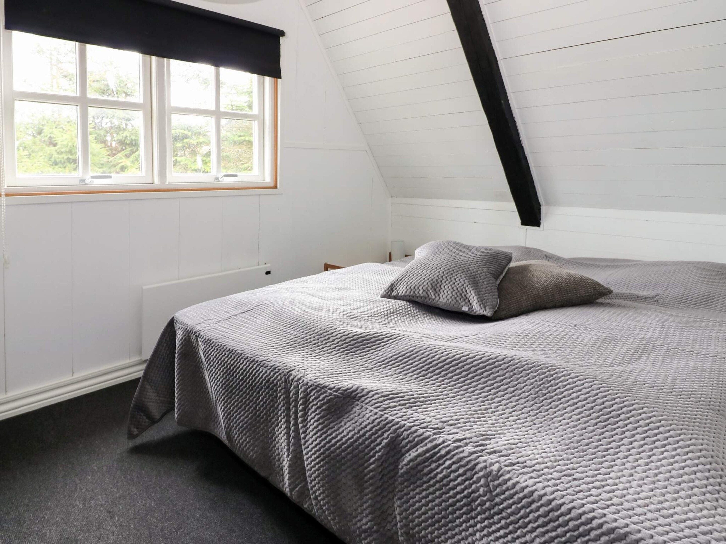 Un dormitorio con techo inclinado, un ventanal grande con cortina negra y una cama con colcha gris, además de un radiador.