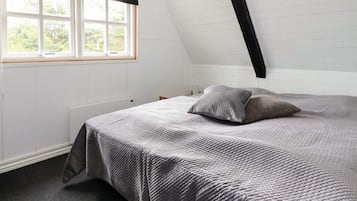 Un dormitorio con techo inclinado, un ventanal grande con cortina negra y una cama con colcha gris, además de un radiador.