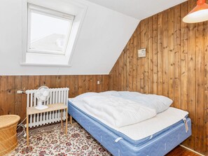 A bedroom with a sloped ceiling, a window, a radiator, a bed, a table, and a lamp.
