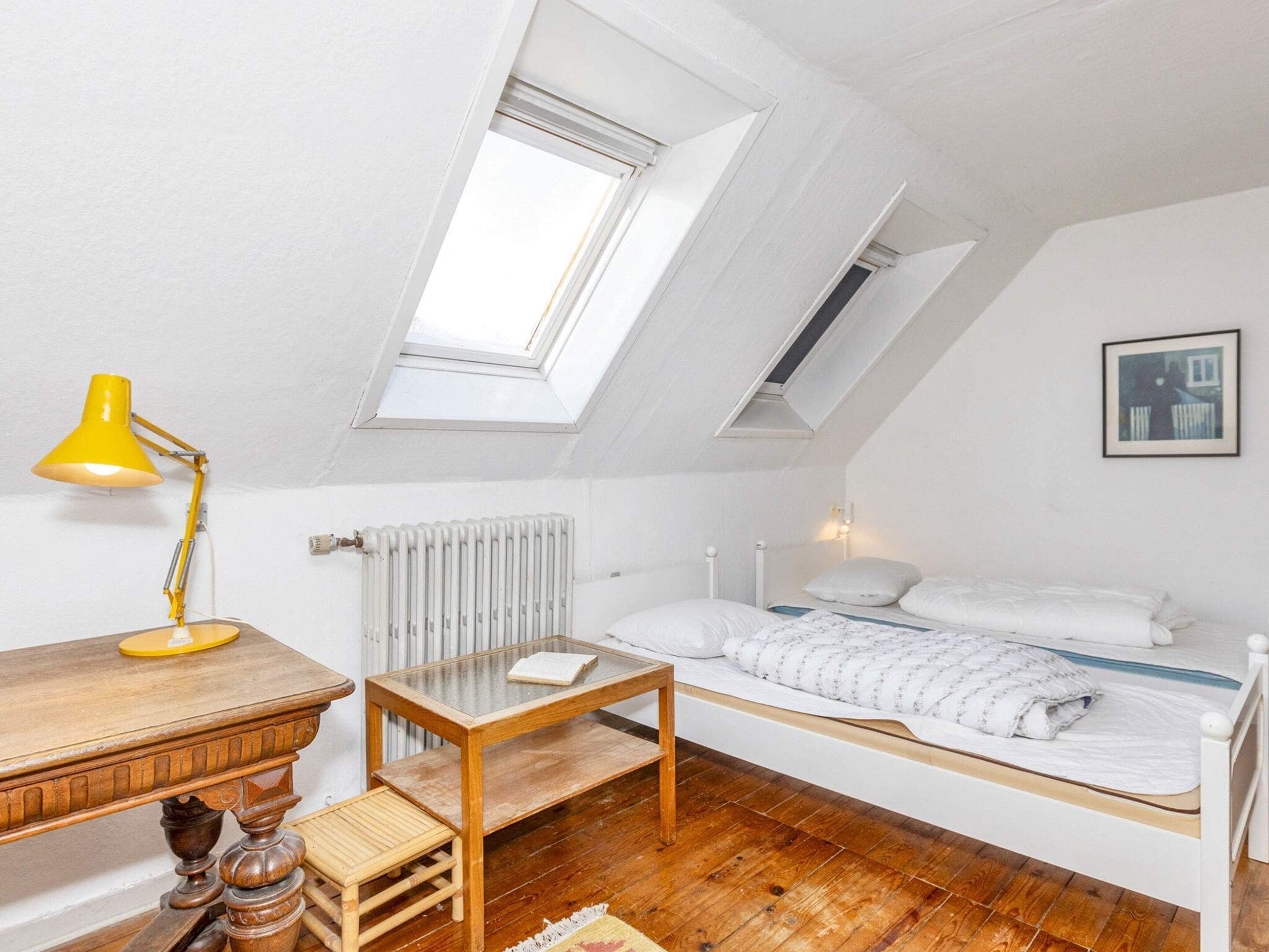 Un dormitorio pequeño y acogedor con techo inclinado, tragaluz, escritorio de madera, una mesita y cama con ropa blanca, además de un radiador.