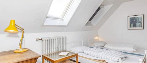A small, cozy bedroom with a sloped ceiling, a skylight, a wooden desk, a small table, a bed with white bedding, and a radiator.
