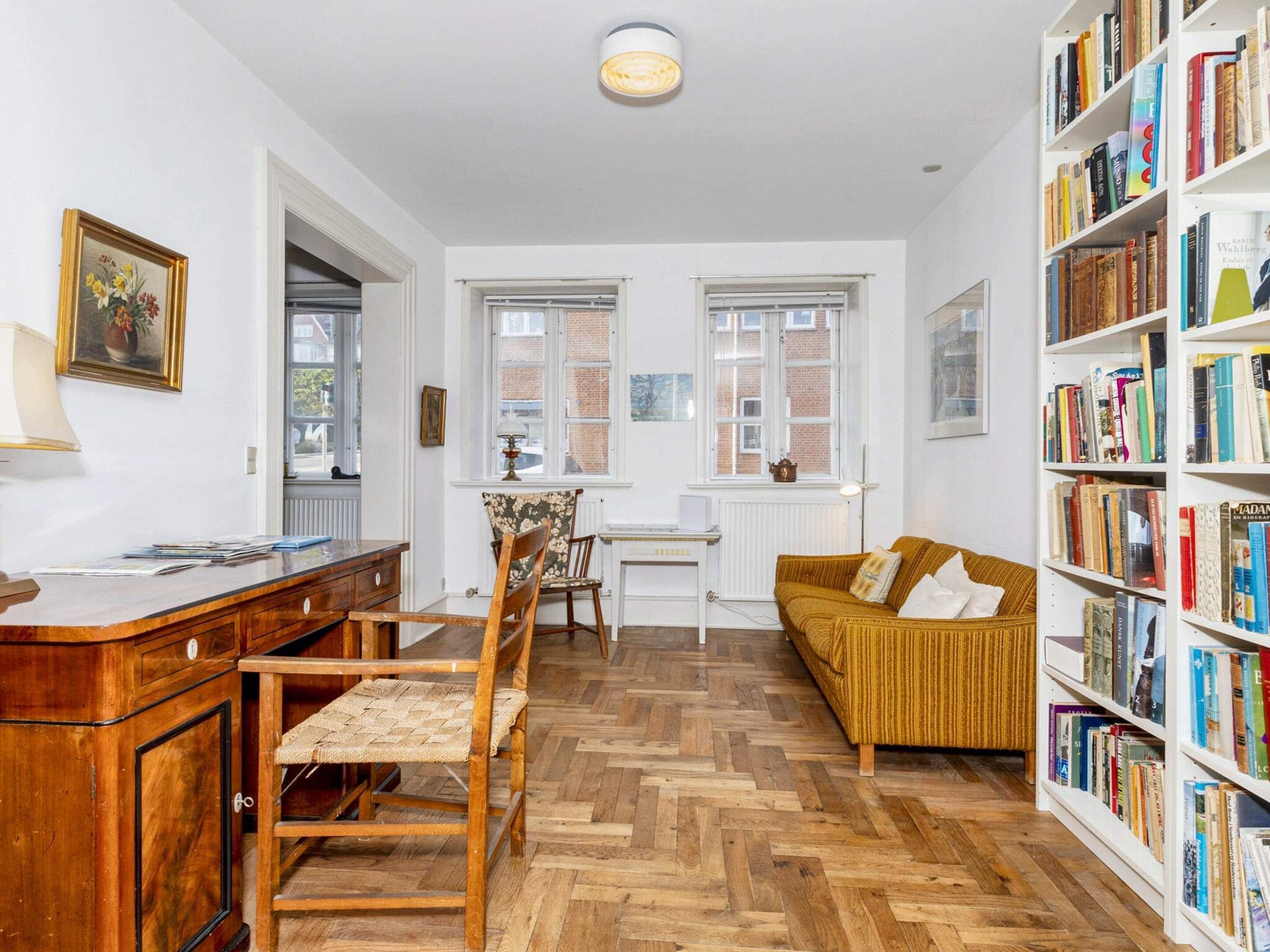 Una habitación con un escritorio de madera, una silla, un sofá, una estantería y una ventana con vistas a edificios.