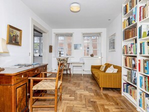 A room with a wooden desk, a chair, a sofa, a bookshelf, and a window with a view of buildings.