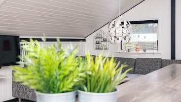 A modern living room with a wooden ceiling, a grey sofa, and a dining table. There are two white planters with green plants in the foreground.