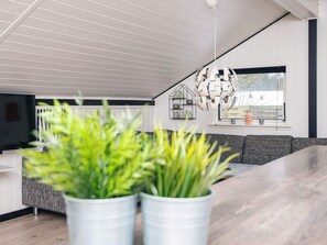 A modern living room with a wooden ceiling, a grey sofa, and a dining table. There are two white planters with green plants in the foreground.