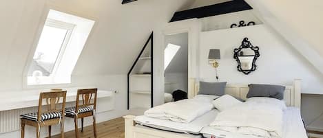 A bedroom with a sloped ceiling, a bed, a desk with chairs, a mirror, and a wall-mounted lamp.