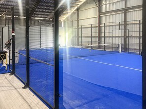 Een overdekte tennisbaan met een blauwe ondergrond, omringd door metalen hekken en een net in het midden.