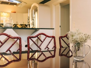 A dining area with a glass table and three wicker chairs. A clear glass pitcher with flowers is on the table.
