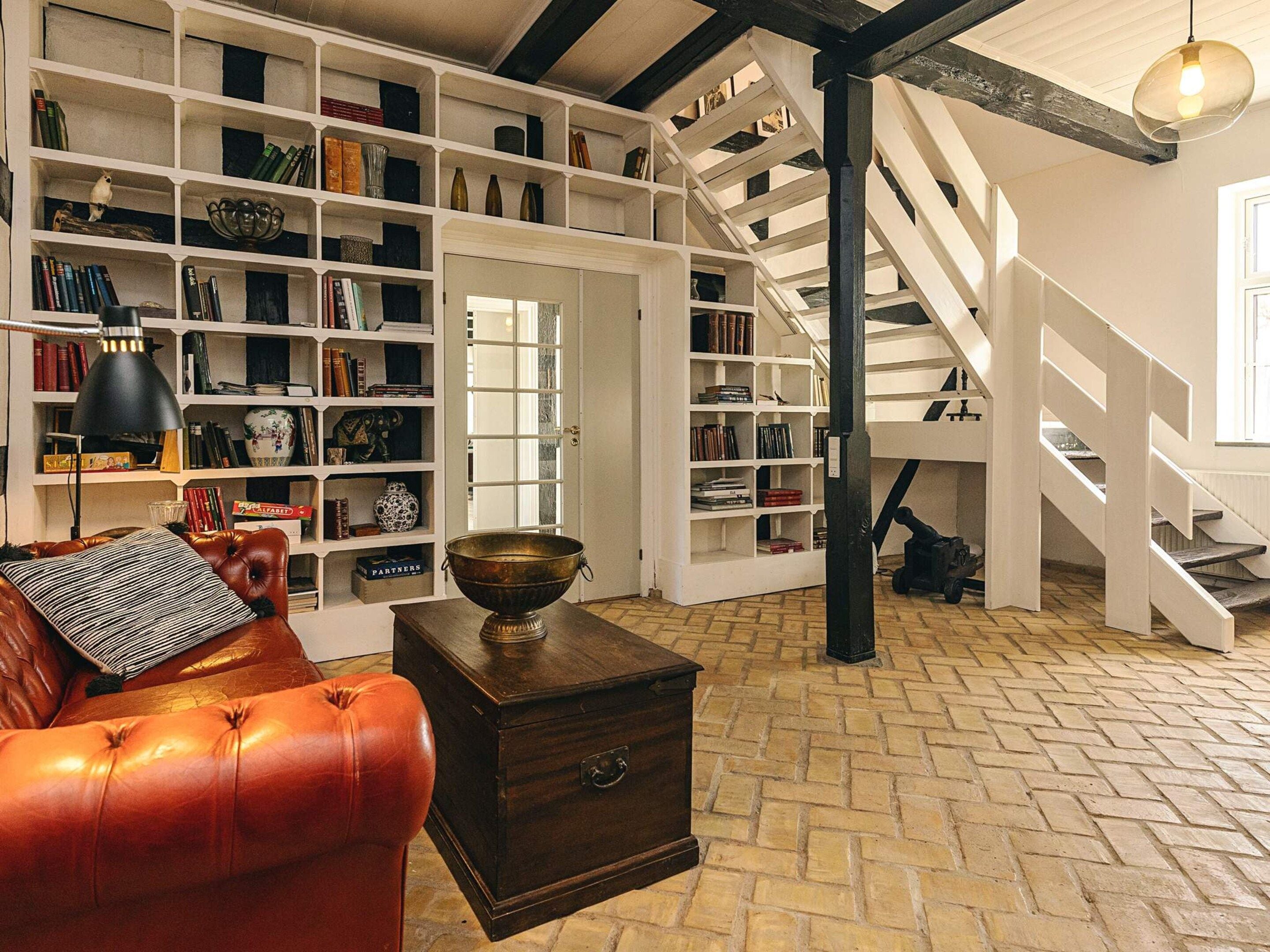 A spacious room with a leather sofa, a wooden chest, and a large bookshelf filled with books and decorative items. A staircase with a wooden railing leads to an upper level.