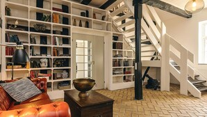 A spacious room with a leather sofa, a wooden chest, and a large bookshelf filled with books and decorative items. A staircase with a wooden railing leads to an upper level. - Holiday Home in Svendborg (Svendborg)