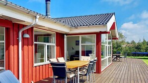 A red building with a wooden deck, a dining area with chairs and a table, and a large window.