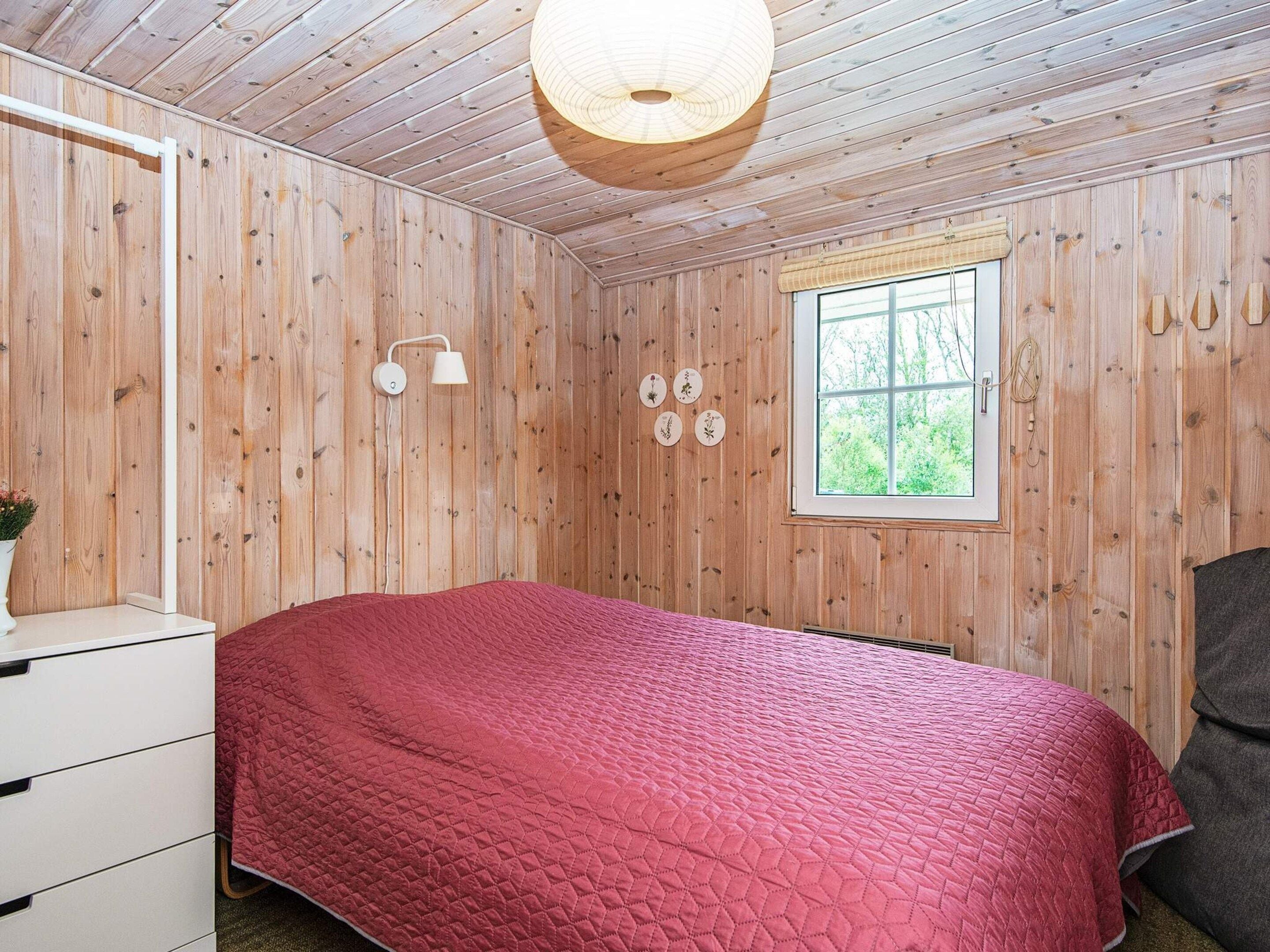 A bedroom with a wooden ceiling, a bed with a red quilt, a bedside table, a lamp, and a window with a view of greenery.