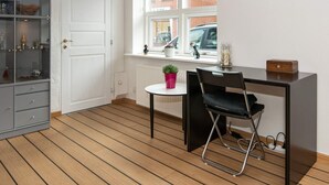 Une pièce avec un plancher en bois, une porte blanche, une fenêtre donnant sur une voiture et des immeubles, un bureau avec une chaise et un meuble avec des portes en verre.