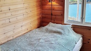 A small, cozy room with wooden walls, a single bed with a patterned blanket, a bedside lamp, and a window offering a view of houses and water. - 6 Person Holiday Home in Austrheim (Austrheim)