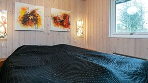 Een slaapkamer met houten wanden, een groot bed met een zwart sprei, twee abstracte schilderijen, een raam met uitzicht op bomen en een wandlamp.