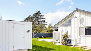 A white RV with a small porch and a satellite dish, set in a grassy backyard with a blue fence and trees in the background.