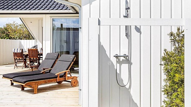 A wooden deck with two lounge chairs and a table, a white building with a tiled roof, and a glass door with a shower.