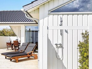 A wooden deck with two lounge chairs and a table, a white building with a tiled roof, and a glass door with a shower.