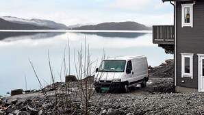 A white van parked by a house with a view of a calm lake and mountains in the background.