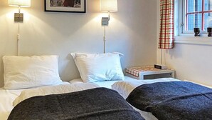 Een tweepersoonsbed met wit beddengoed en twee zwarte plaids, een nachtkastje met een lamp, een ingelijst schilderij aan de muur en een raam met een geruit gordijn.