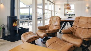 A modern interior with a wood-burning stove, leather chairs, a dining table, and large windows. - Holiday Home in LÃ¸kken (Lokken)