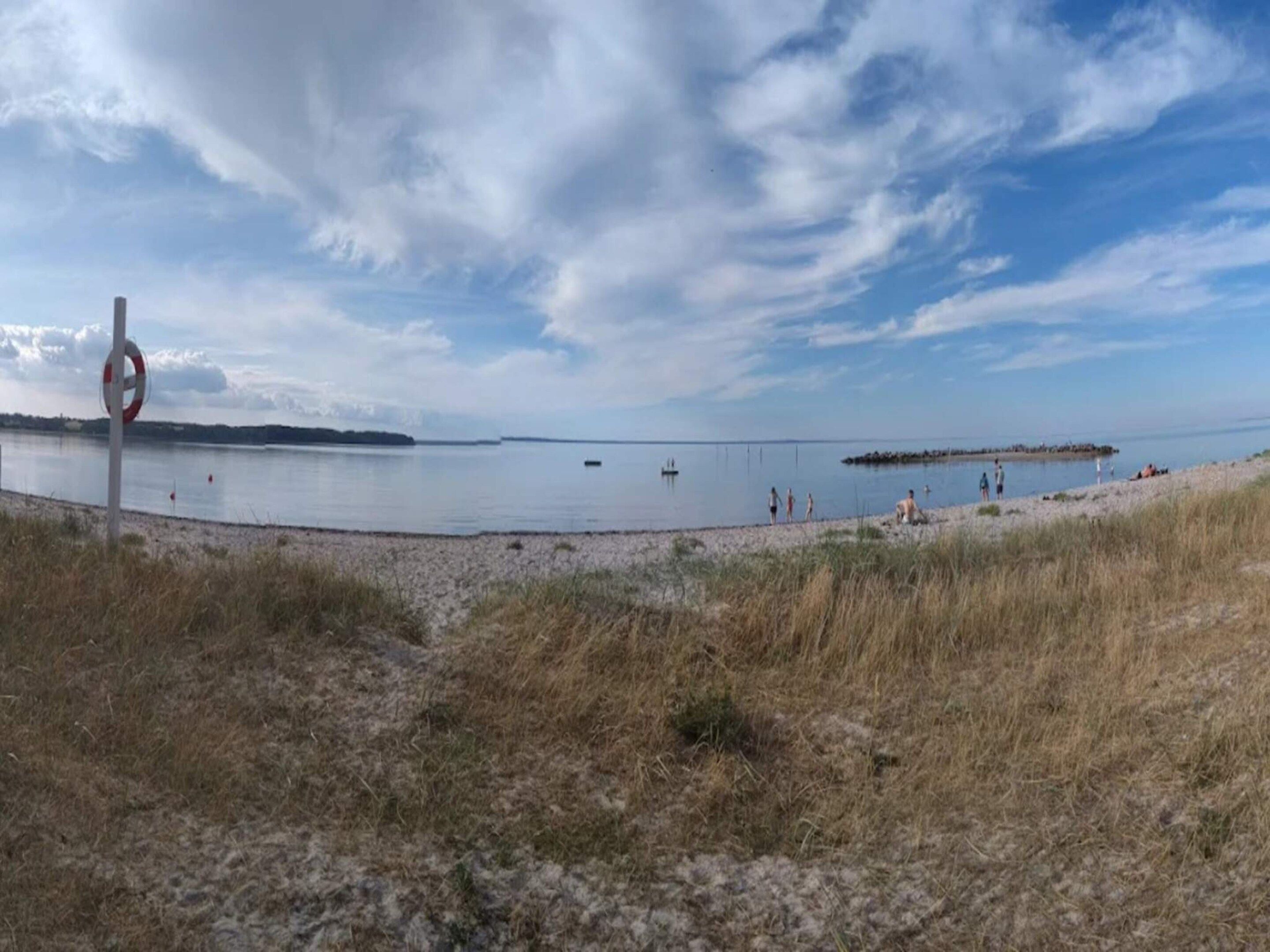 Ein Strand mit einem Rettungsbojen-Schild, ruhigem Wasser und einer entfernten Küstenlinie.
