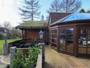A wooden cabin with a glass conservatory, stone pillars, and a patio area with outdoor furniture.