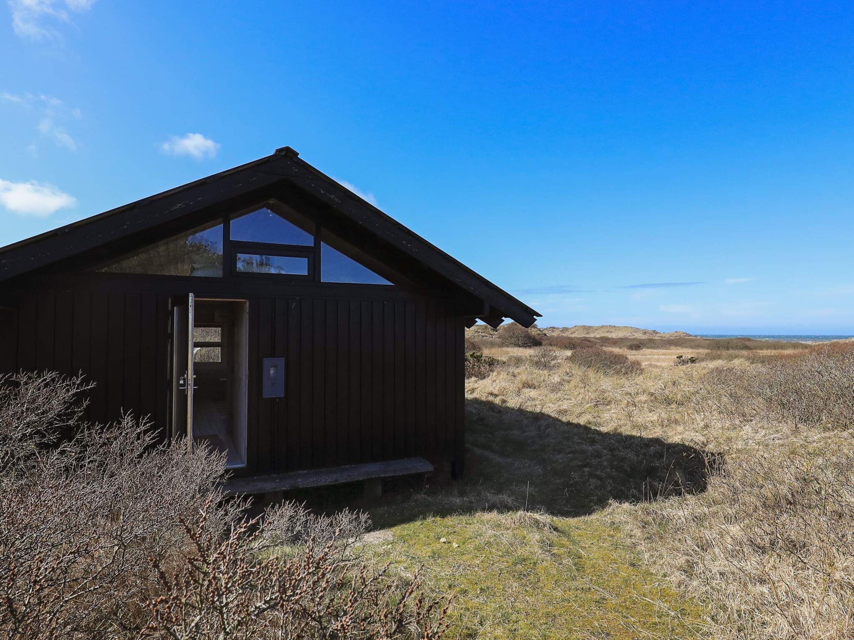 Una cabana de fusta amb una finestra gran i una porta, envoltada d'herba seca i arbustos.