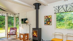A cozy room with a wood-burning stove, a comfortable chair, a small table, and a large window with a view of greenery. - 5 Person Holiday Home in Ry (Ry)